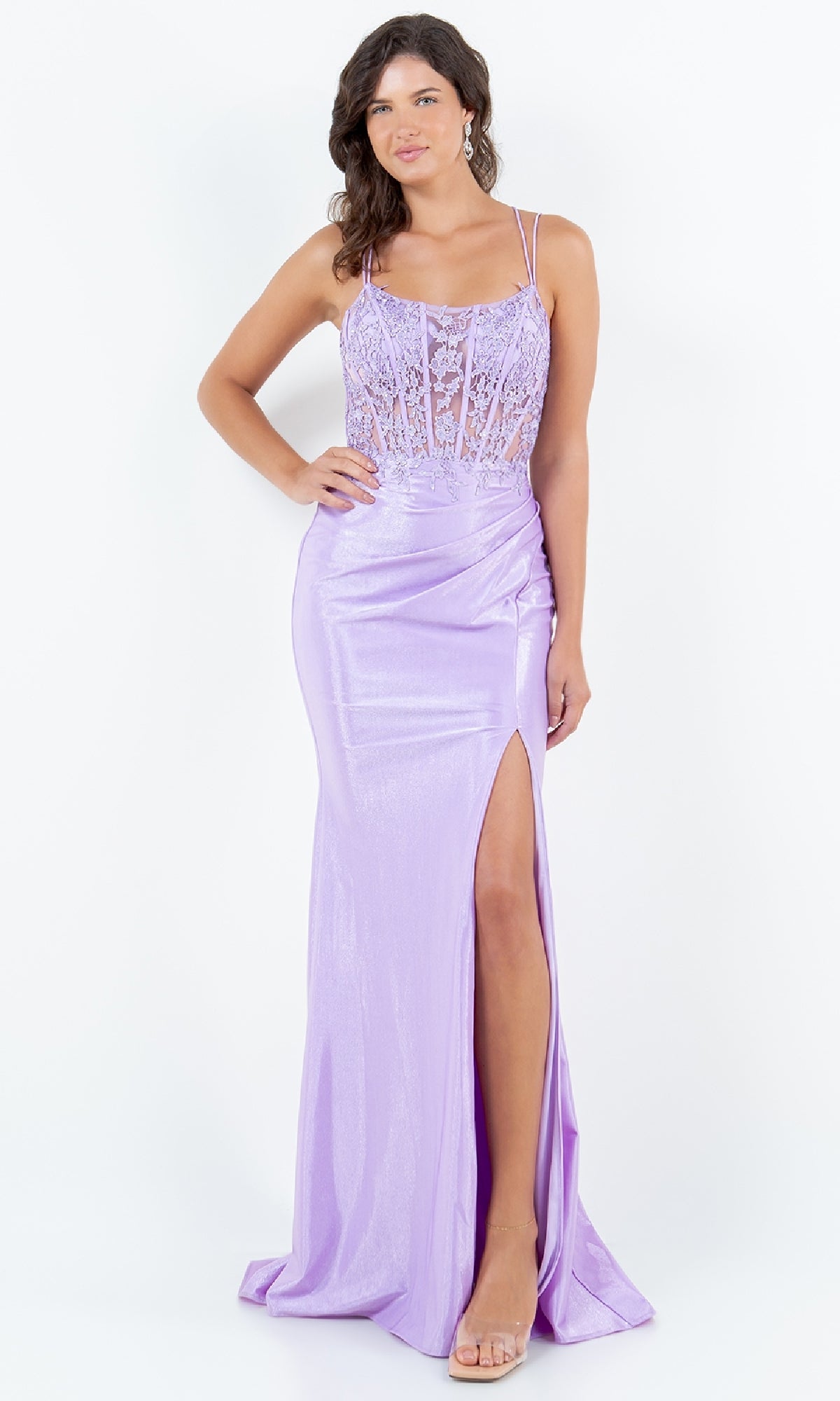 A woman wearing the Lace-Up Lilac Prom Gown (Cinderella Couture 8185J), a floor-length, shimmery purple dress with a lace bodice, thin straps, and a high slit, stands against a white background.
